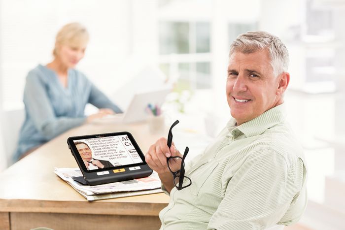 An aging couple is seated at a kitchen table where a man is reading a newspaper with the help of the exploré 8 video magnifier. At the same time, a woman uses a personal computer.